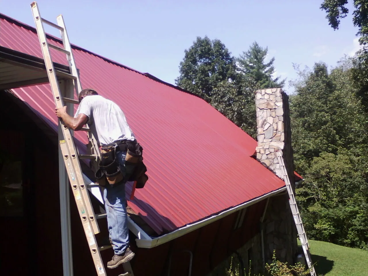 Roofer on a ladder beside a red metal roof and stone chimney — Soffit & Fascia Repair by Roofing Brothers in Kalispell